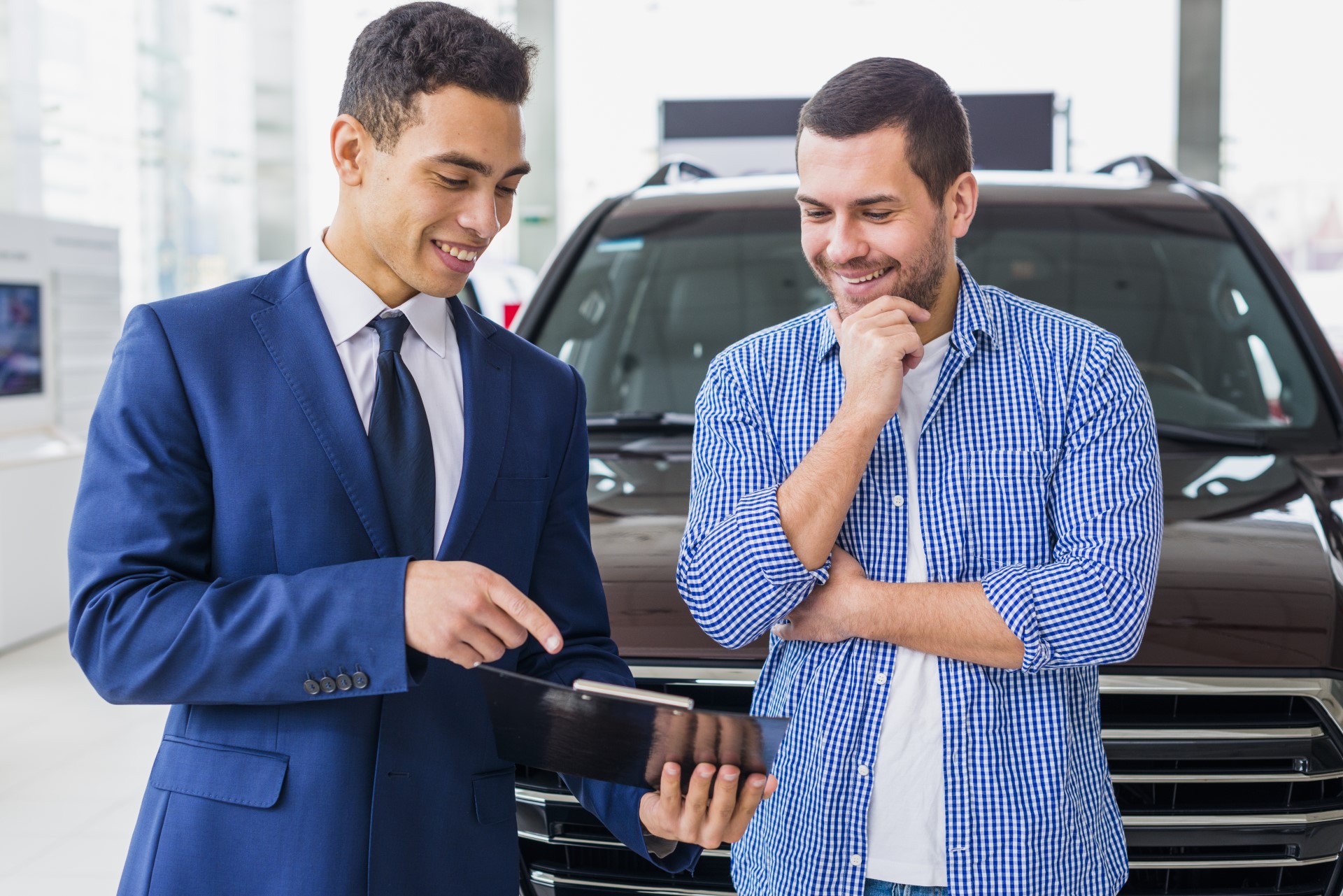 A car salesman in a blue suit shows a tablet to a customer in a checkered shirt, discussing a vehicle in a bright dealership.